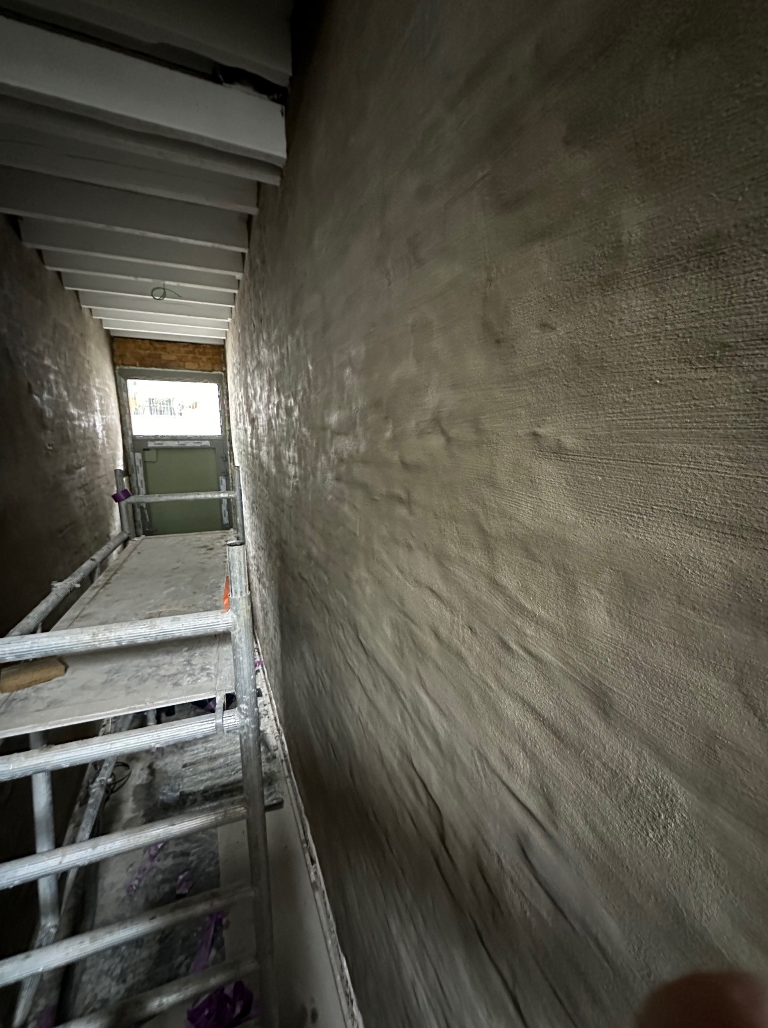 Wąskie wnętrze w trakcie prac wykończeniowych z nałożoną szarą warstwą na ścianach, widoczne rusztowanie i okno na końcu korytarza.