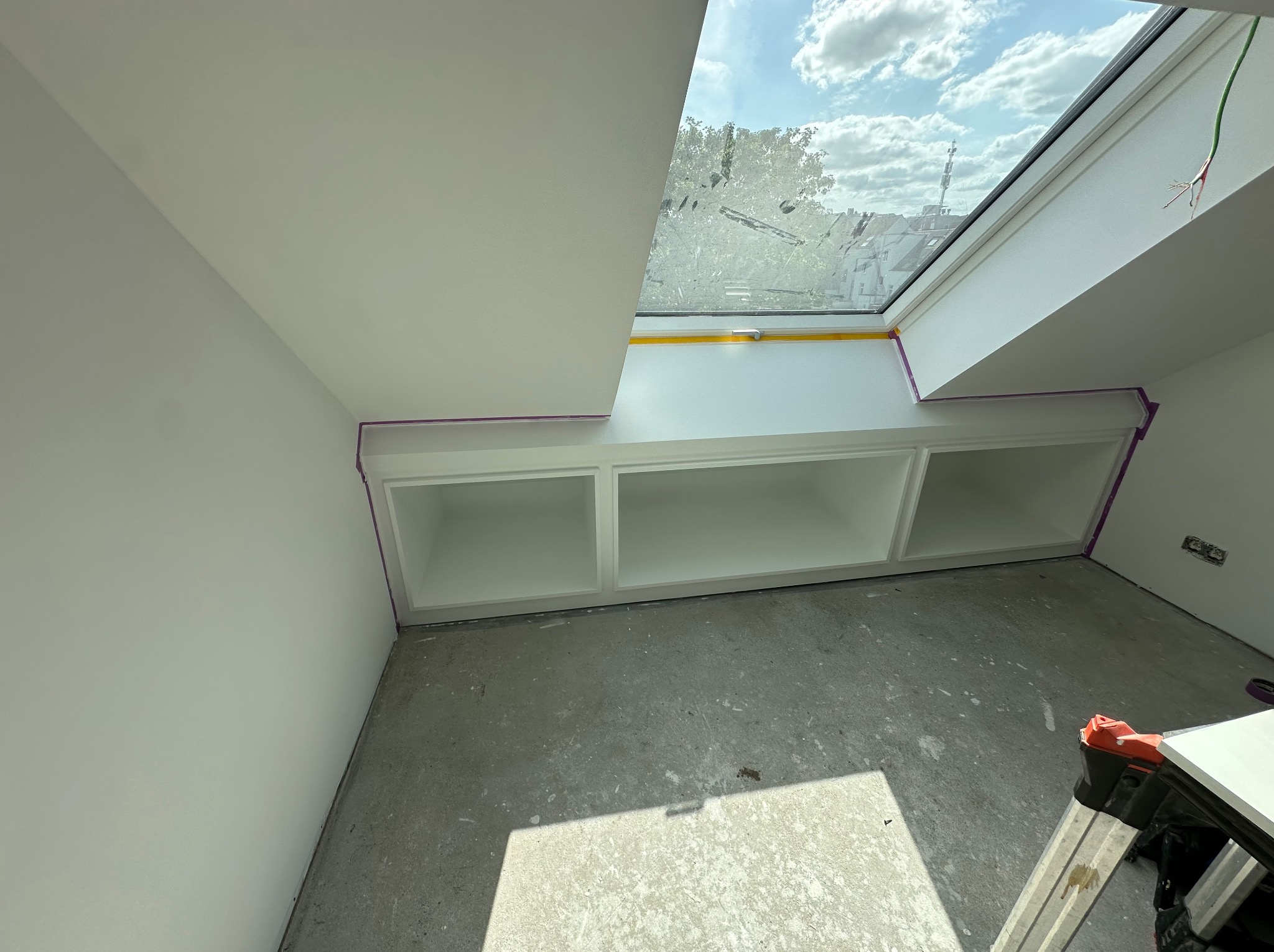Wnętrze poddasza z oknem dachowym i wnęką z trzema półkami, świeżo pomalowane na biało, zabezpieczone taśmą malarską, surowy beton na podłodze, drabina w rogu.