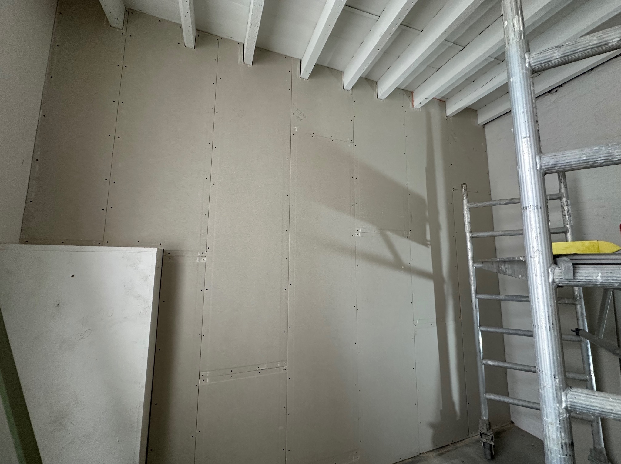 Ściana pokryta płytami karton gips, widoczne wkręty, białe belki stropowe i aluminiowa drabina, etap prac wykończeniowych.