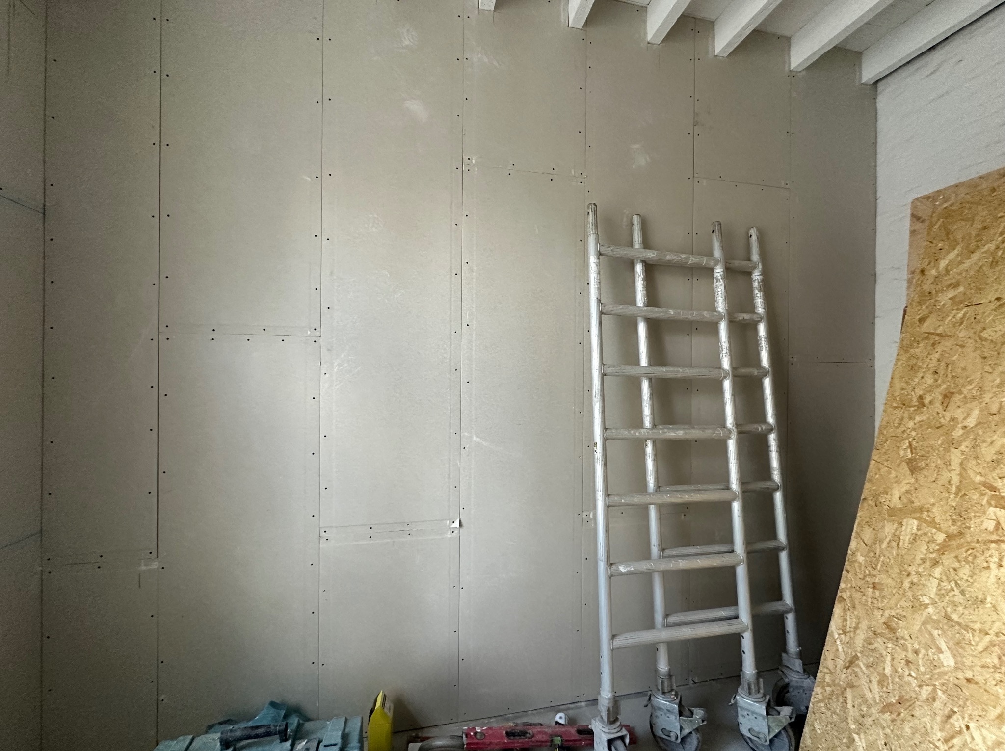 Ściana pokryta płytami gipsowo-kartonowymi, widoczne łączenia i wkręty, oparta o nią aluminiowa drabina na kółkach, w tle fragment płyty OSB.
