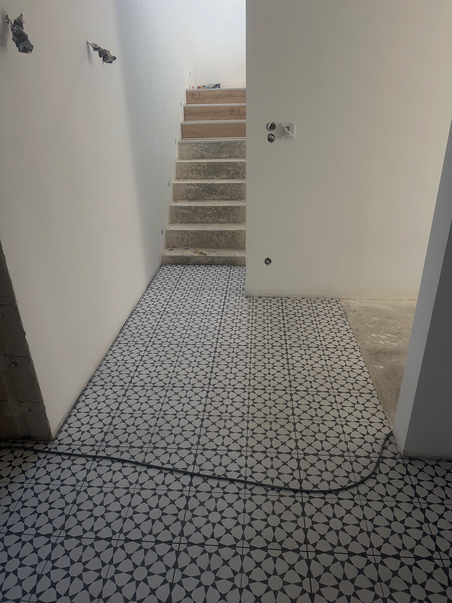 Ułożona, wzorzysta posadzka w przedpokoju z betonowymi schodami i gładkimi, białymi ścianami. Widoczne niedokończone prace na ścianach i podłodze.