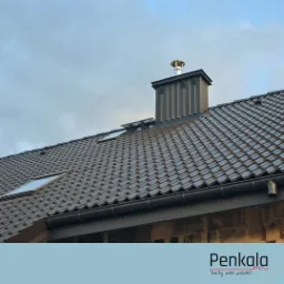 Nowy, brązowy dach z dachówki ceramicznej z lukarnami i metalowym kominem z nasadą, widok z dołu na tle pochmurnego nieba.