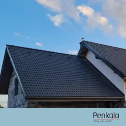 Czarny dach z dachówki ceramicznej na domu w trakcie budowy, widoczna surowa ściana z pustaków i fragment niebieskiego nieba z chmurami. W dolnej części logo firmy dekarskiej.