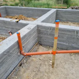 Surowe fundamenty domu z widocznymi instalacjami kanalizacyjnymi, betonowe ściany z odciśniętą fakturą szalunku, wystające pręty zbrojeniowe i fragment narzędzi budowlanych.