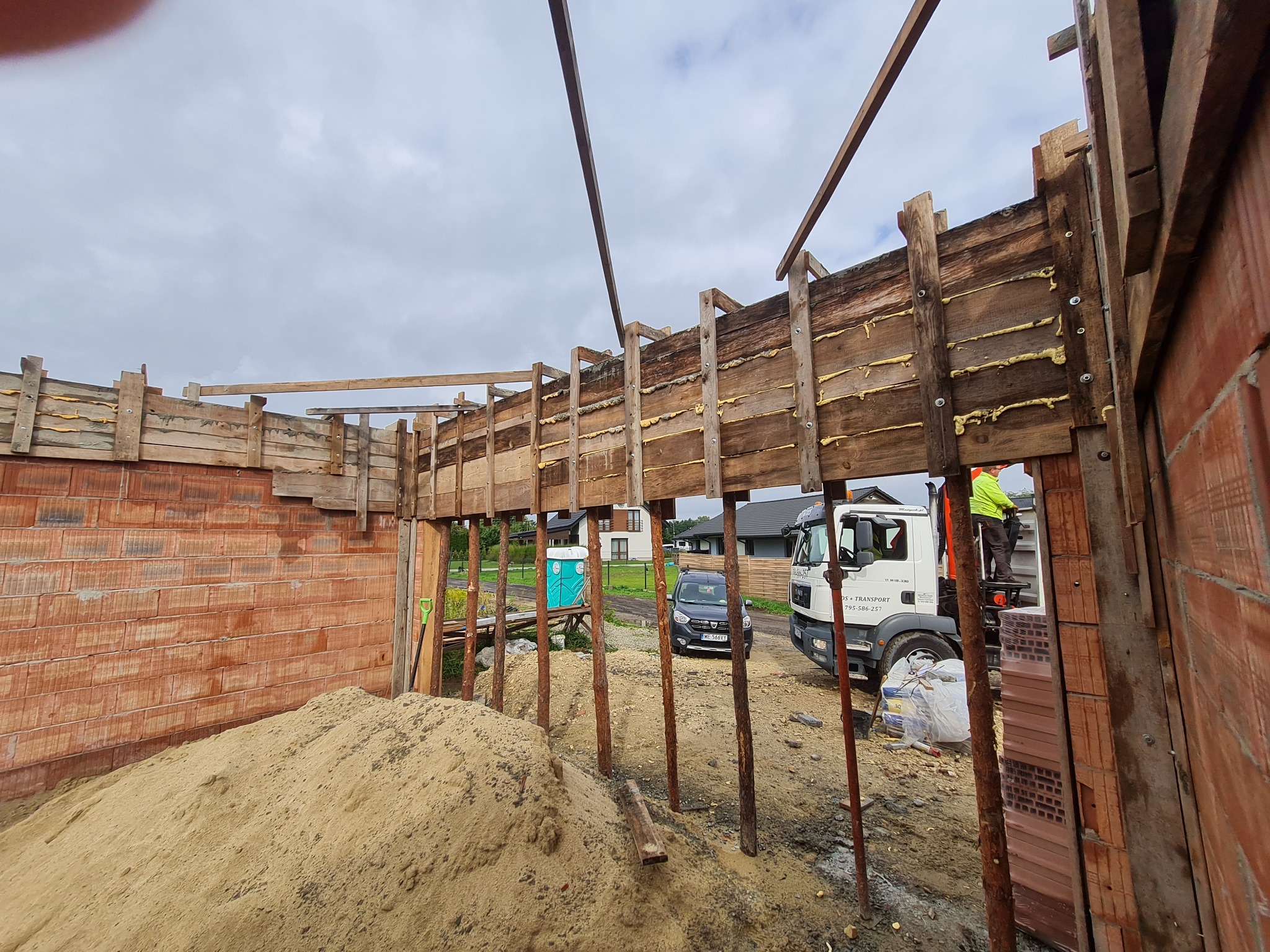 Wznoszenie ścian domu z cegły ceramicznej, widoczne drewniane szalunki wypełnione pianką montażową, stos cegieł, ciężarówka na budowie i pracownik w żółtej kamizelce.