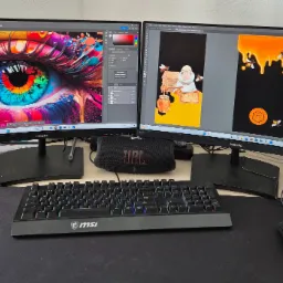 Stanowisko pracy grafika z dwoma monitorami prezentującymi projekty: abstrakcyjne, kolorowe oko oraz ilustracja pszczelarza i miodu. Widoczna klawiatura MSI, głośnik JBL i myszka.