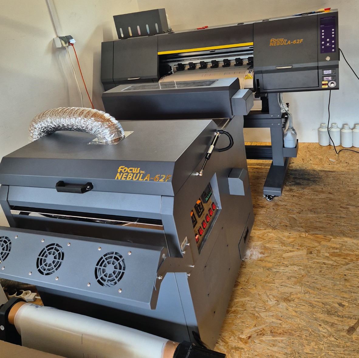 Dwa urządzenia do druku DTF: drukarka wielkoformatowa Focus Nebula-62F oraz urządzenie do utrwalania nadruku z widocznym wężem wentylacyjnym i rolką folii.