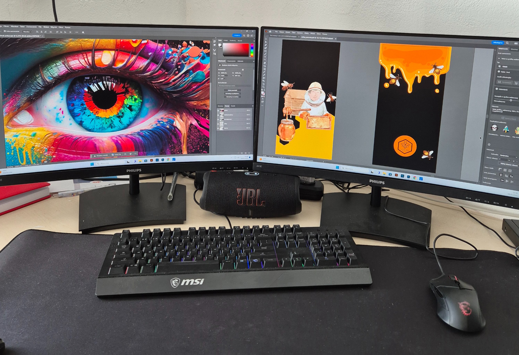 Stanowisko pracy grafika z dwoma monitorami prezentującymi projekty: abstrakcyjne, kolorowe oko oraz ilustracja pszczelarza i miodu. Widoczna klawiatura MSI, głośnik JBL i myszka.