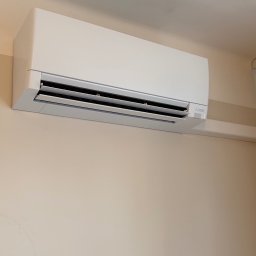 Soplo Klimatyzacja - Biały klimatyzator Mitsubishi Electric zamontowany na beżowej ścianie w pomieszczeniu. Widoczne żaluzje i diody sygnalizacyjne.