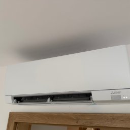 Soplo Klimatyzacja - Biała klimatyzacja Mitsubishi Electric zamontowana na ścianie, widoczna dolna część ramy drzwi z drewna. Minimalistyczny design, nowoczesne wnętrze.