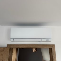 Soplo Klimatyzacja - Biała jednostka klimatyzacji Mitsubishi Electric zamontowana na ścianie nad drewnianą futryną drzwiową w jasnym wnętrzu. Widoczny fragment sufitu.