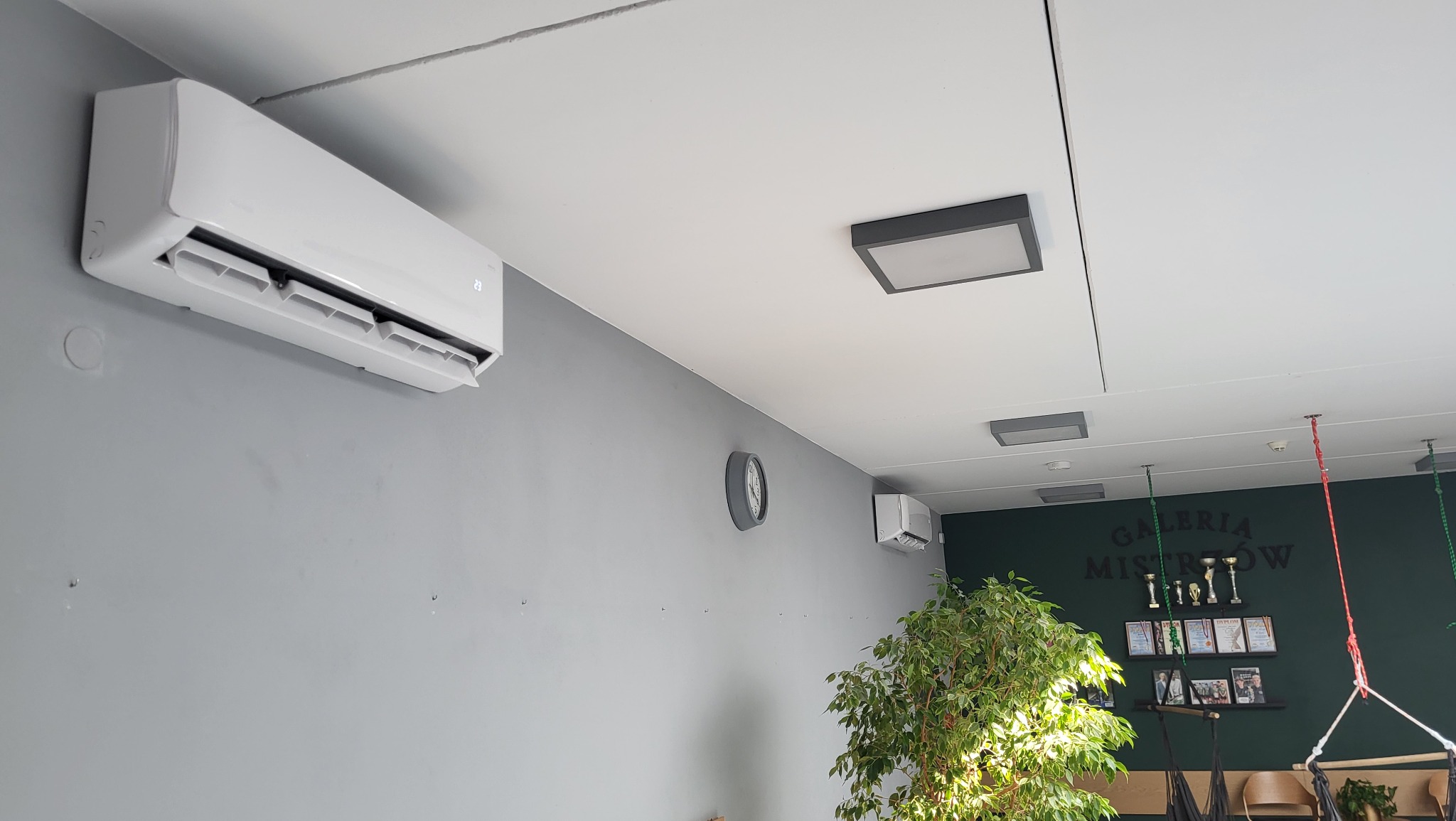 Biały klimatyzator ścienny w pomieszczeniu biurowym z zegarem, rośliną doniczkową i wiszącymi huśtawkami hamakowymi w tle.