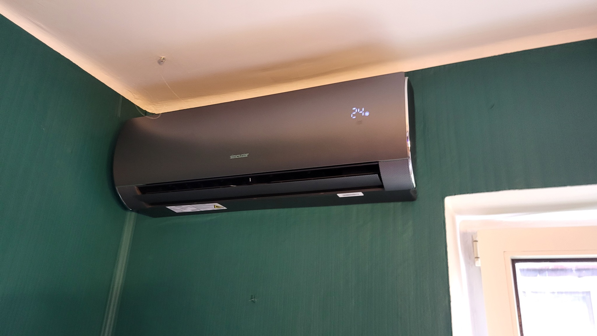 Nowoczesny klimatyzator Sinclair w kolorze grafitowym, zamontowany na zielonej ścianie z widocznym wyświetlaczem temperatury 24 stopnie Celsjusza.