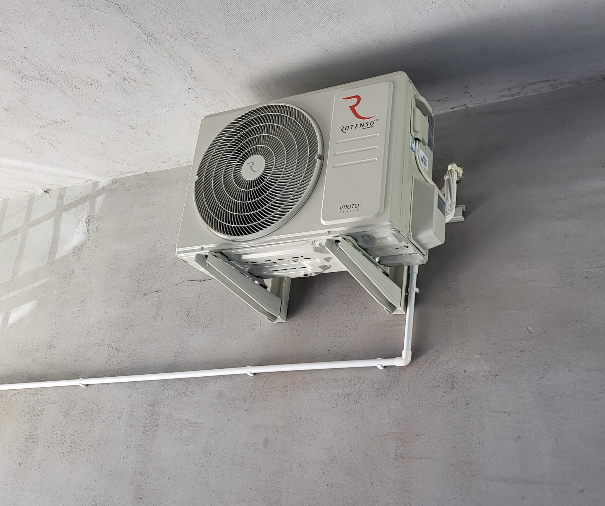 Biała jednostka zewnętrzna klimatyzatora Rotenso Imoto Series zamontowana na szarej, surowej ścianie z widocznymi rurami odprowadzającymi skropliny.