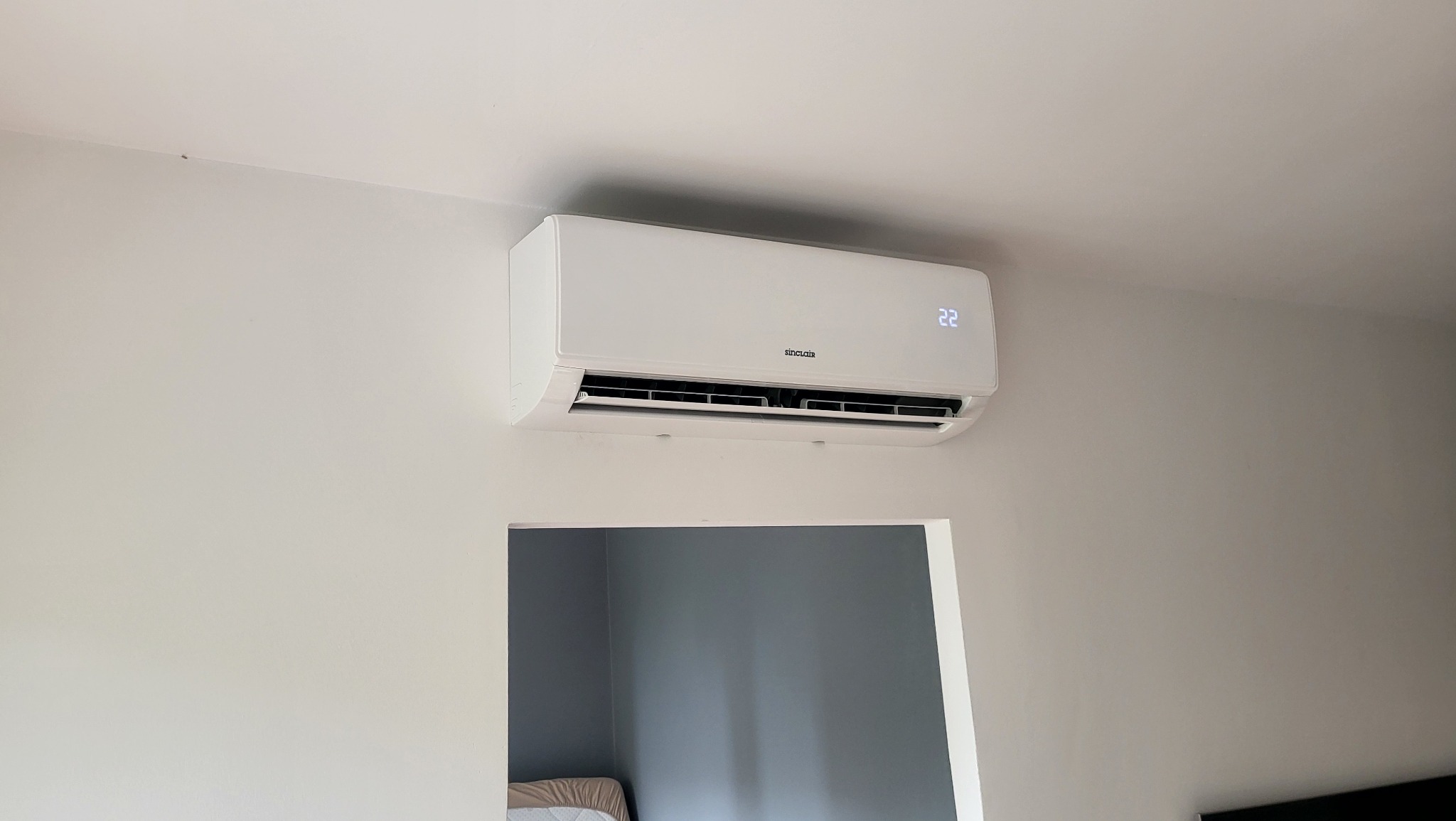 Biały klimatyzator Sinclair zamontowany na jasnoszarej ścianie w pokoju, widoczny wyświetlacz wskazujący temperaturę 22 stopnie Celsjusza, poniżej widoczne fragmenty innego pomieszczenia...