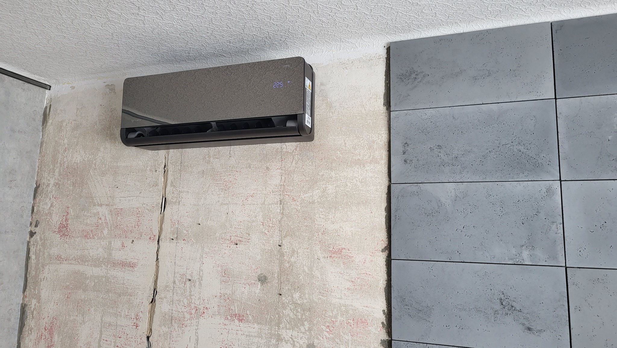 Ciemny klimatyzator na ścianie w trakcie remontu, obok widoczne szare płytki imitujące beton.