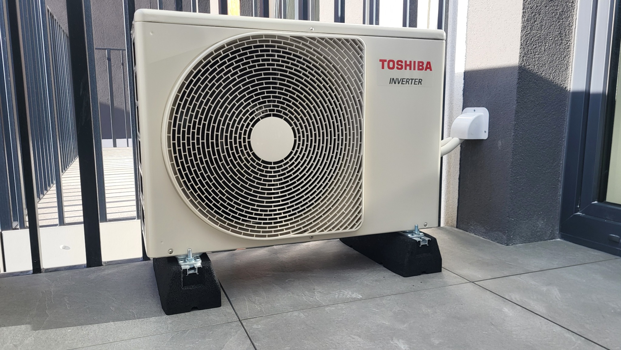Zewnętrzna jednostka klimatyzacji Toshiba Inverter zamontowana na czarnych, gumowych podkładkach antywibracyjnych na szarym, płytkowym balkonie, widoczne metalowe barierki i elewacja budynku.