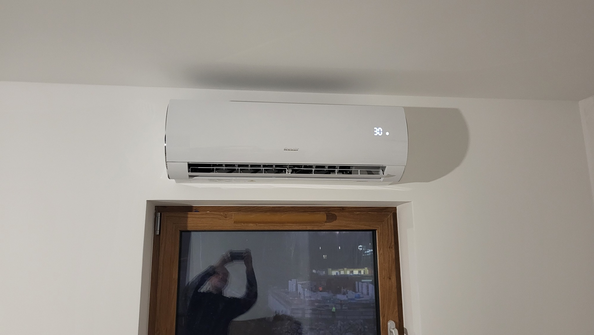 Biały klimatyzator Sinclair zamontowany nad oknem z drewnianą ramą; na wyświetlaczu widoczna temperatura 30 stopni; w oknie odbicie osoby robiącej zdjęcie.