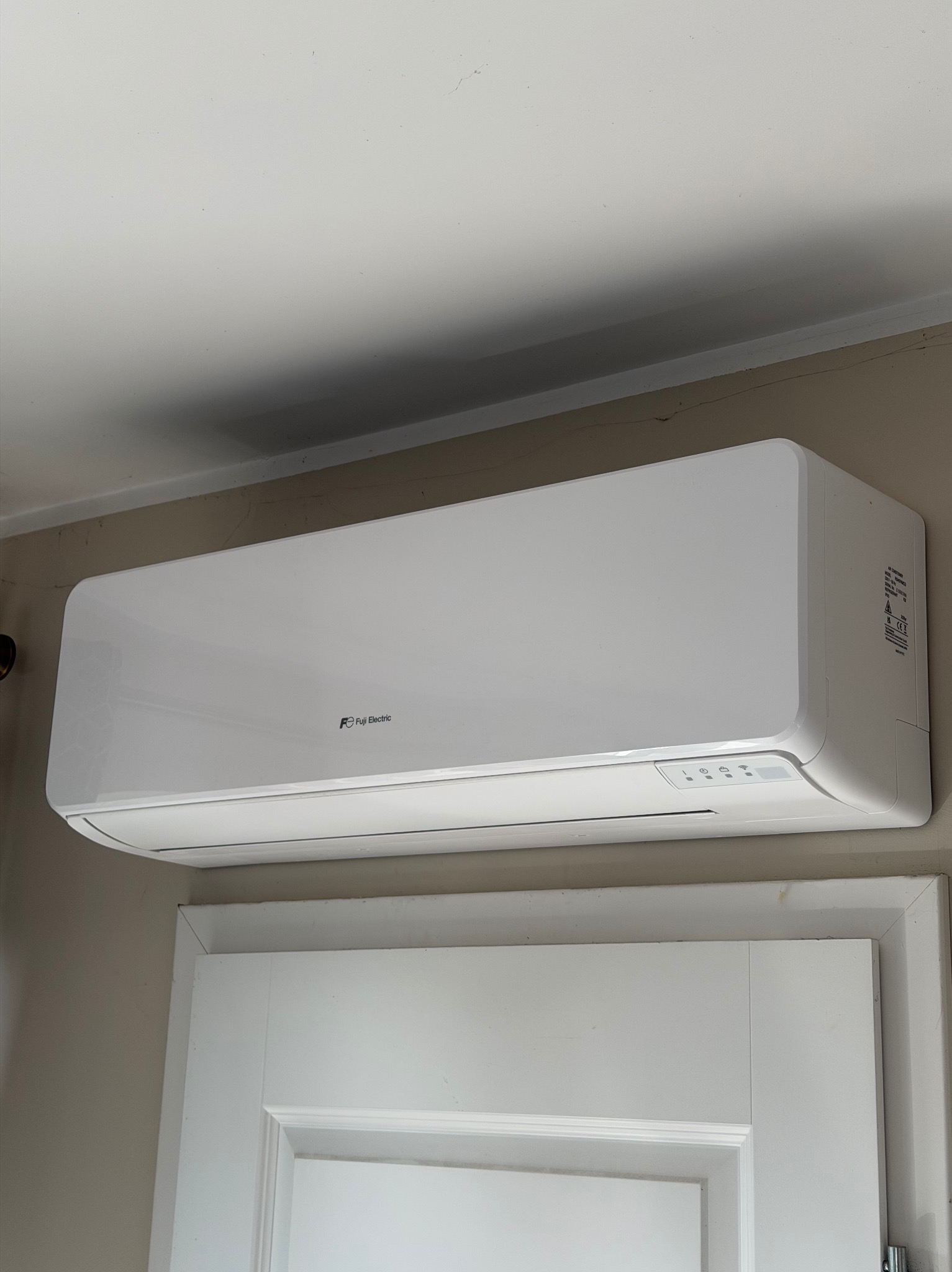 Biała jednostka klimatyzacji Fuji Electric zamontowana nad białymi drzwiami w pomieszczeniu o jasnych ścianach. Widoczne logo producenta i panel sterowania.