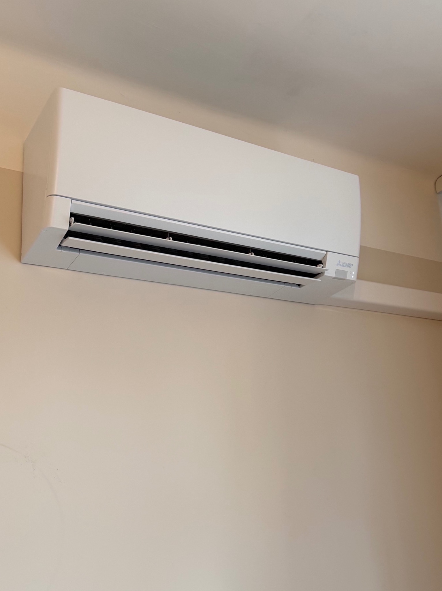 Biały klimatyzator Mitsubishi Electric zamontowany na beżowej ścianie w pomieszczeniu. Widoczne żaluzje i diody sygnalizacyjne.