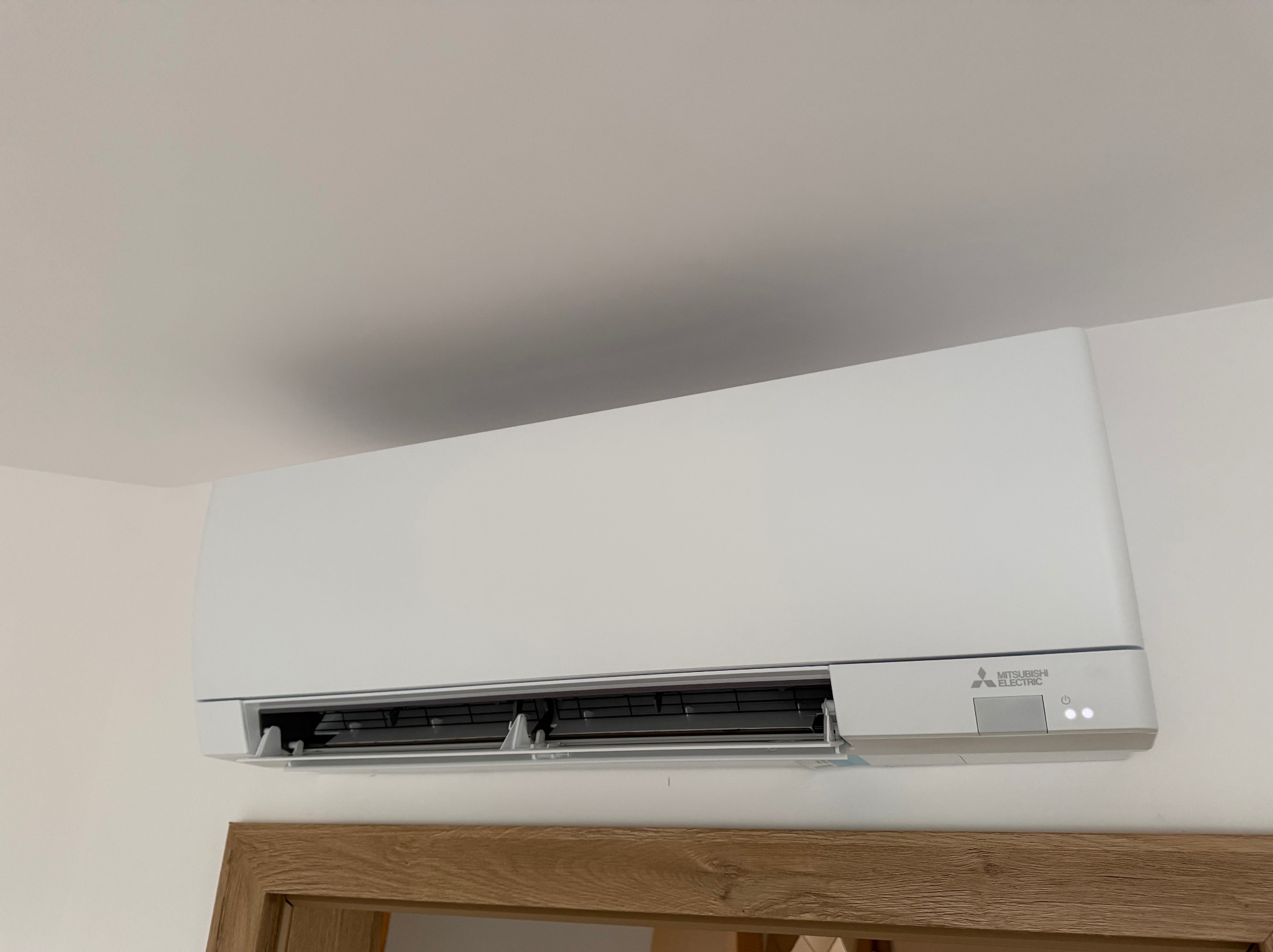 Biała klimatyzacja Mitsubishi Electric zamontowana na ścianie, widoczna dolna część ramy drzwi z drewna. Minimalistyczny design, nowoczesne wnętrze.