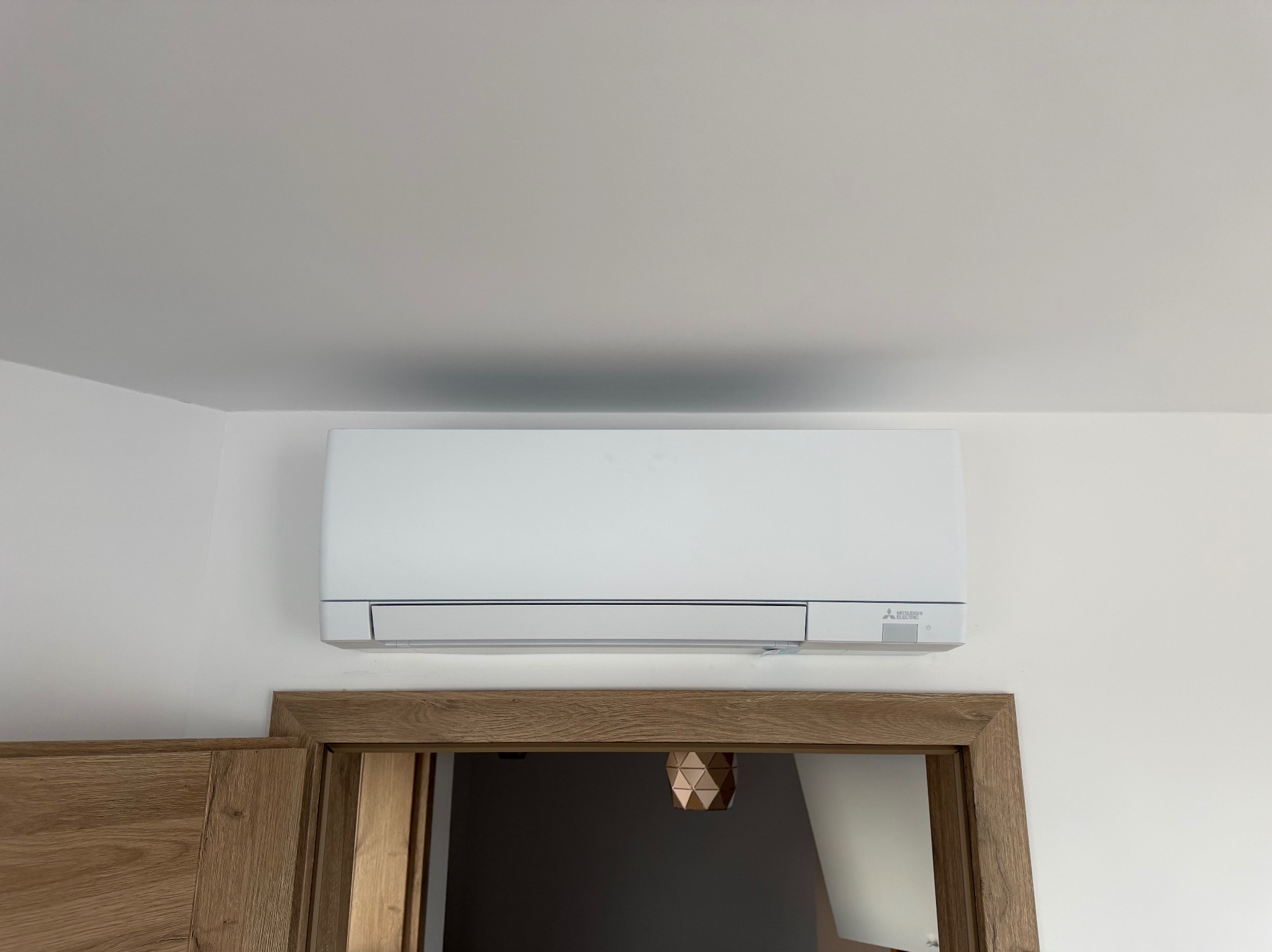 Biała jednostka klimatyzacji Mitsubishi Electric zamontowana na ścianie nad drewnianą futryną drzwiową w jasnym wnętrzu. Widoczny fragment sufitu.