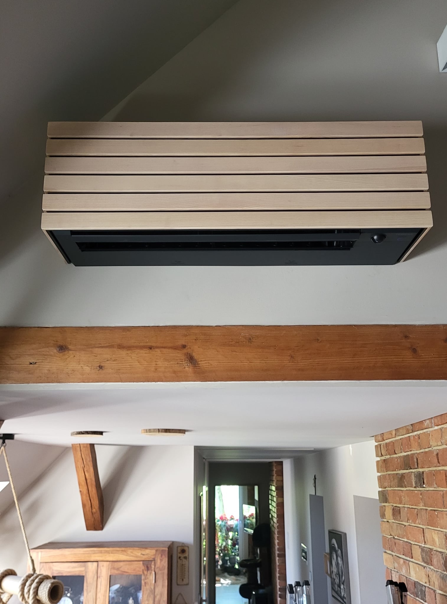 Klimatyzator ścienny z drewnianą obudową w nowoczesnym wnętrzu. Widoczne drewniane belki stropowe i fragment ceglanej ściany. Nietypowe wykończenie klimatyzacji.