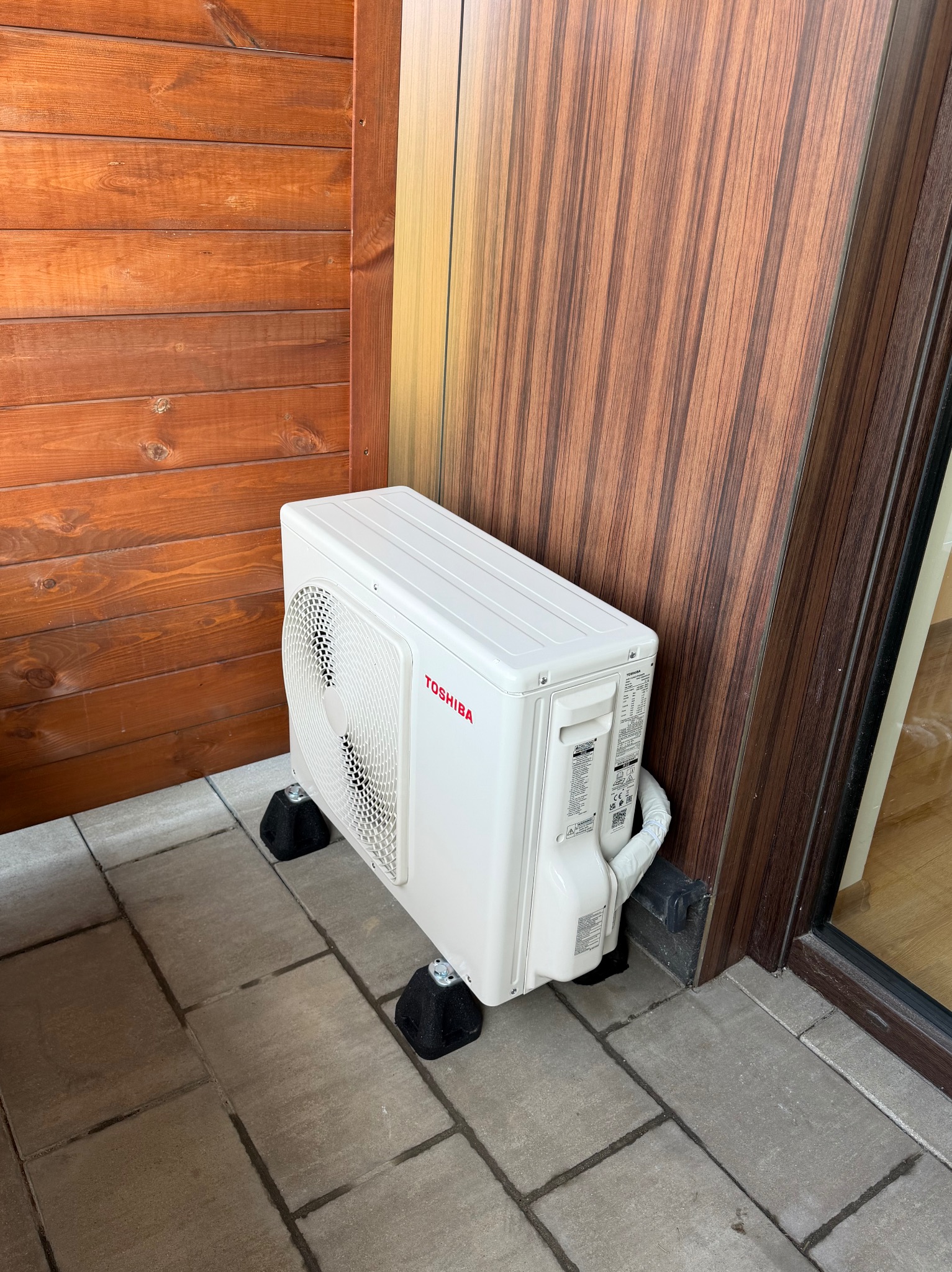 Biała jednostka zewnętrzna klimatyzacji Toshiba, umieszczona na czarnych podkładkach, stoi na szarym bruku przy drewnianej elewacji budynku. Widoczny fragment okna.