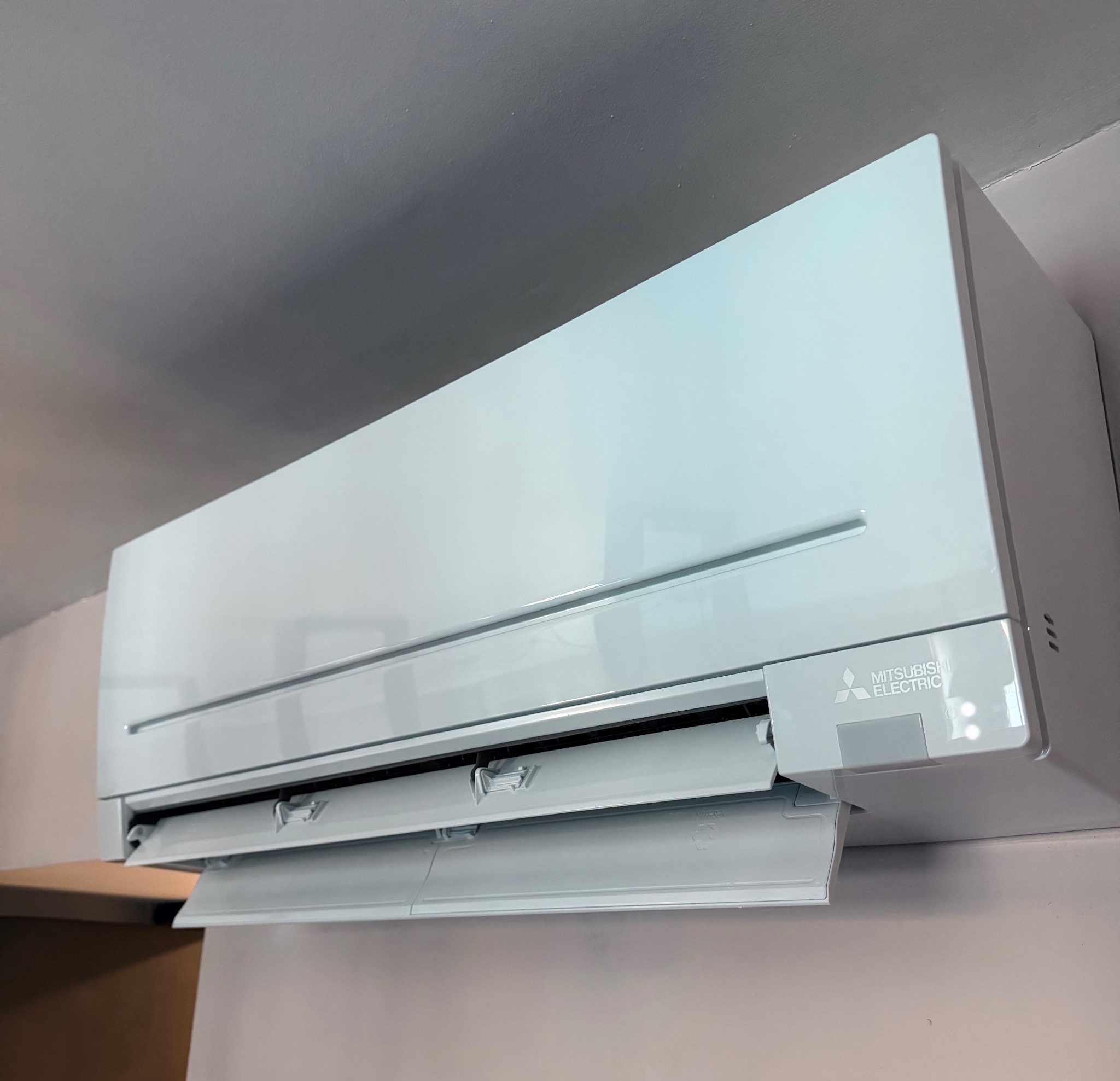 Biała, naścienna jednostka klimatyzacji Mitsubishi Electric z otwartymi klapami, zamontowana w pomieszczeniu o jasnych ścianach i suficie. Widoczne logo producenta.