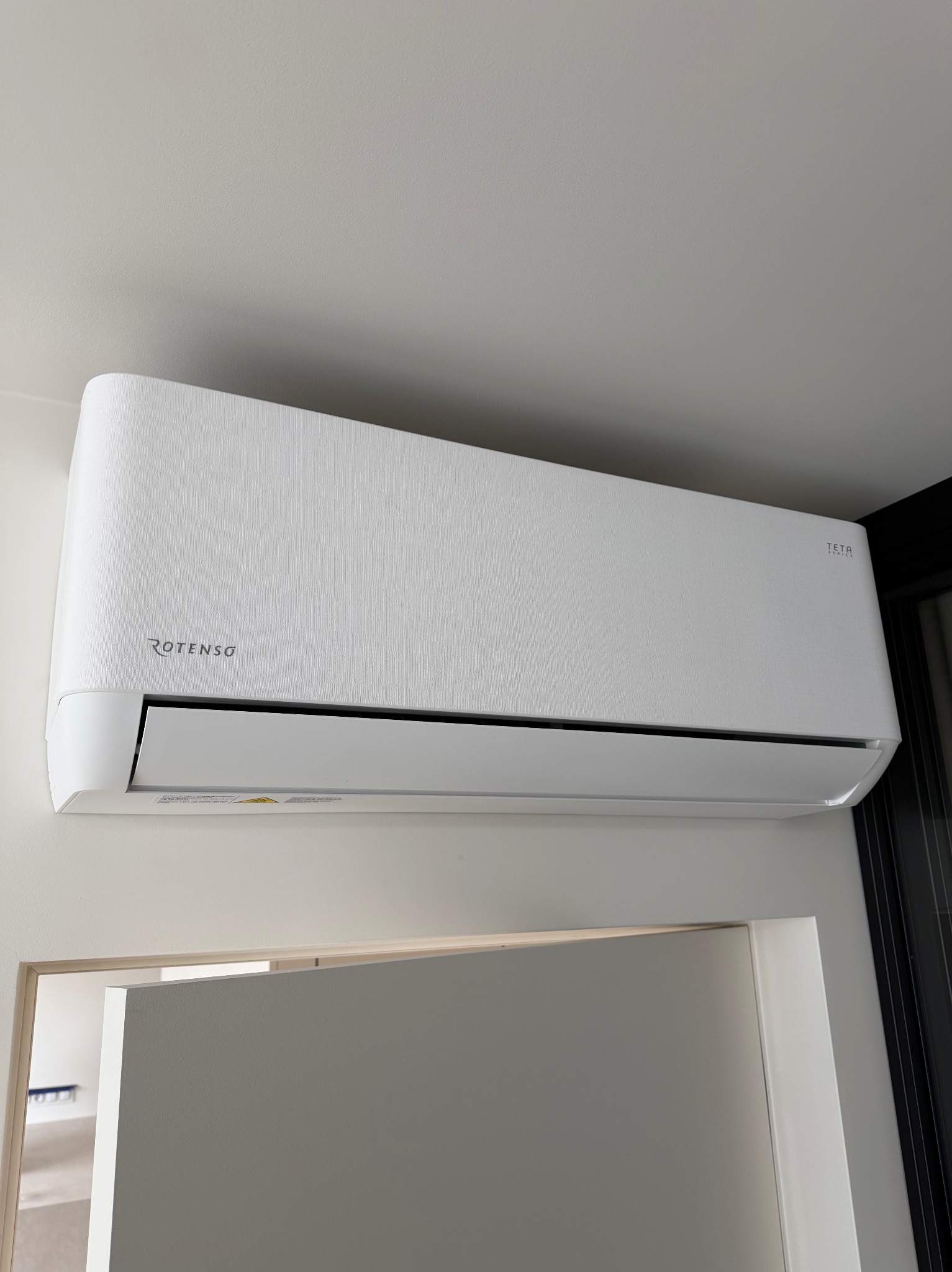Biała klimatyzacja Rotenso Teta zamontowana na ścianie w pomieszczeniu, widoczny fragment drzwi i okna. Minimalistyczny design, jasne wnętrze.