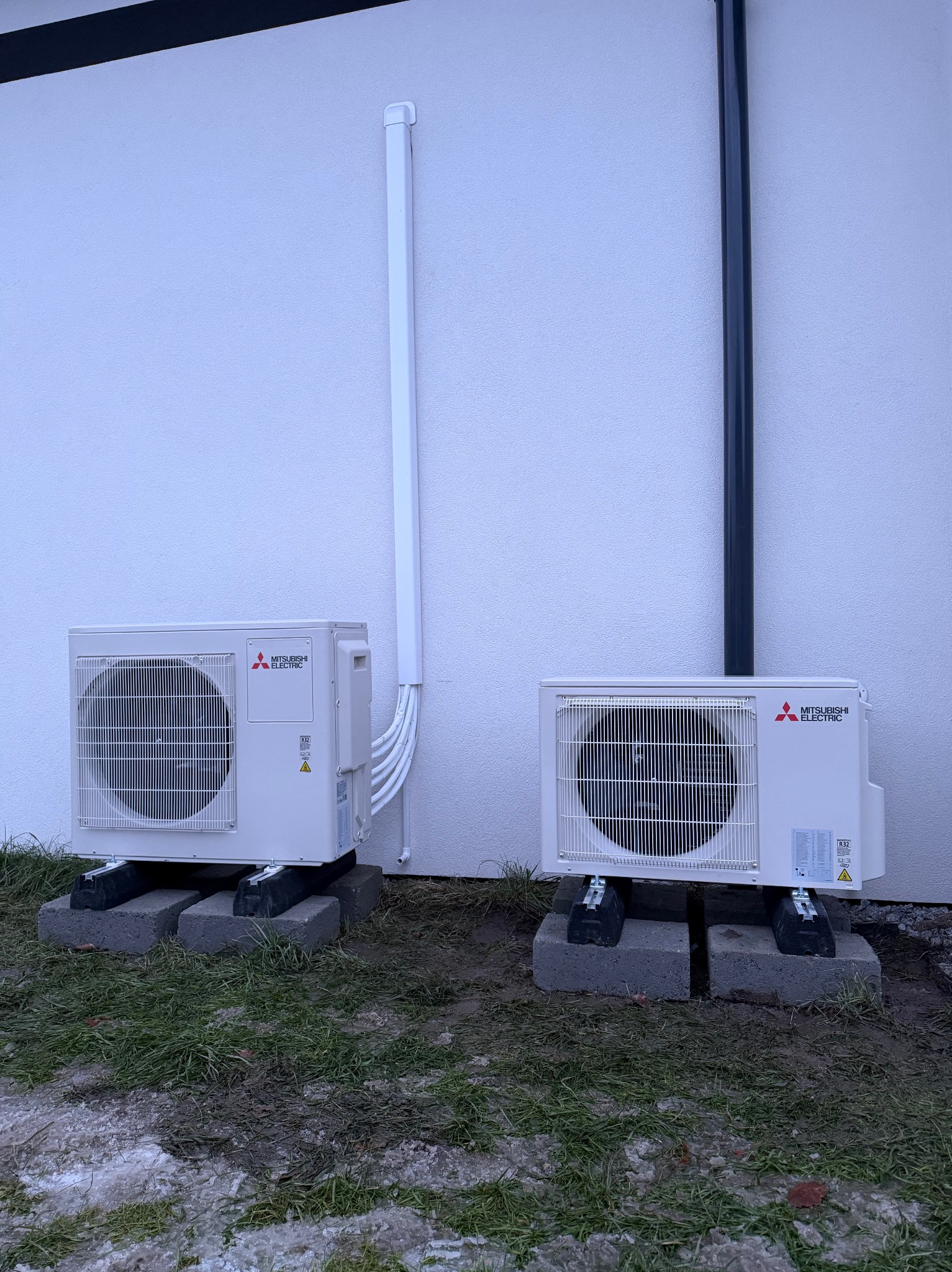 Dwa białe klimatyzatory Mitsubishi Electric zamontowane na betonowych podstawach przy białej ścianie budynku. Widoczne rury odprowadzające. Ujęcie z poziomu gruntu.