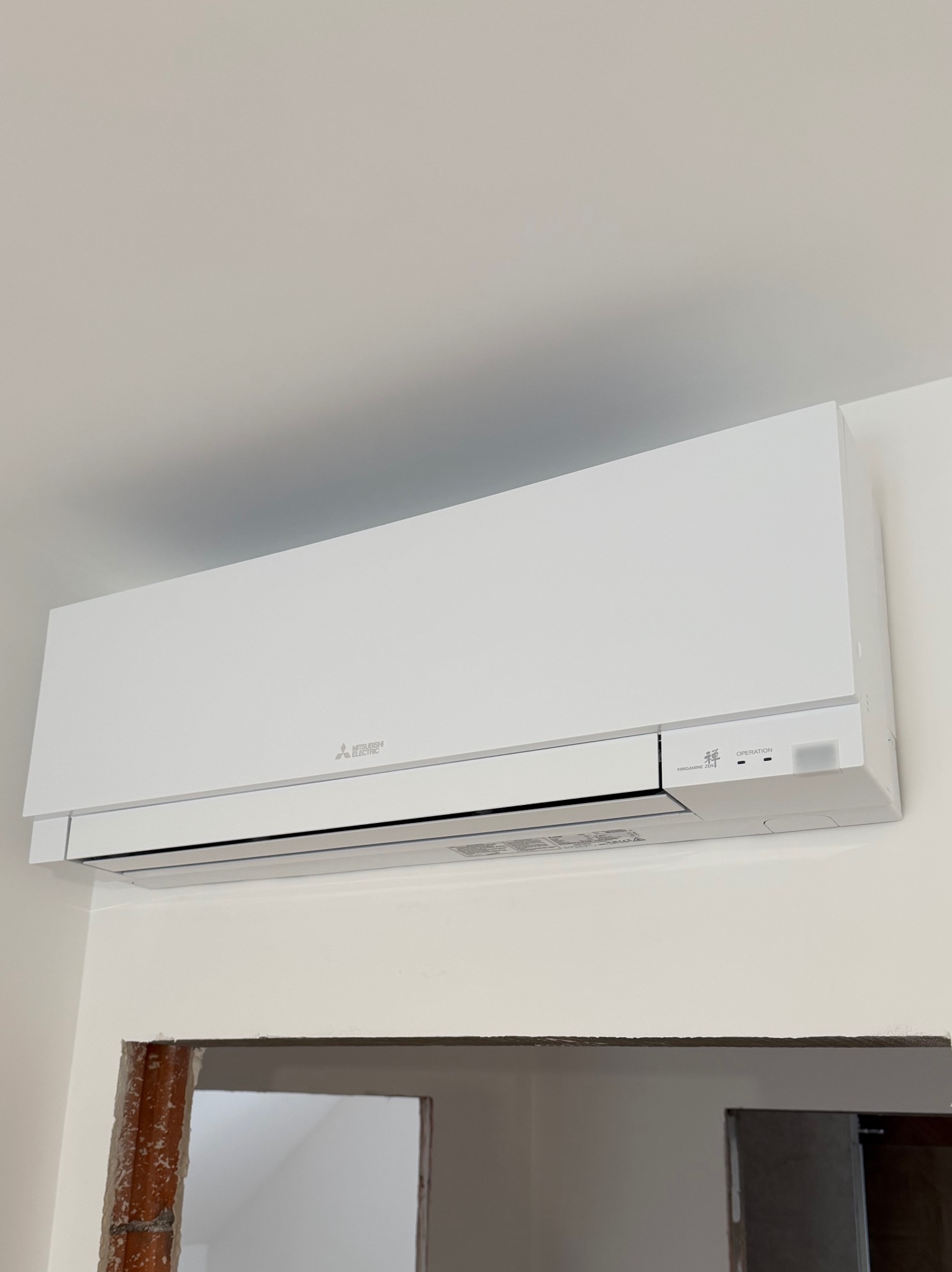 Biała jednostka klimatyzacji Mitsubishi Electric, model Kirigamine Zen, zamontowana na ścianie nad otworem drzwiowym w trakcie remontu.