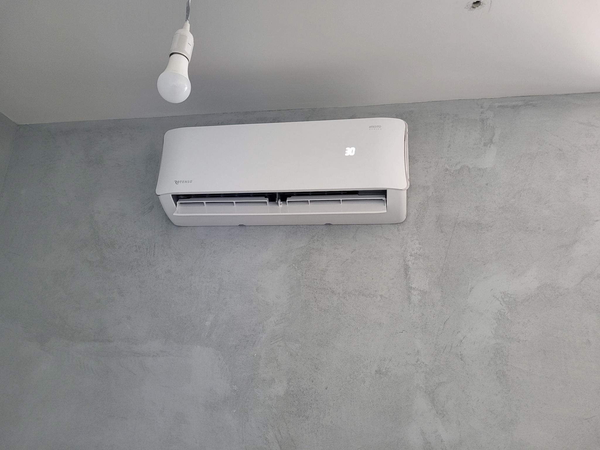 Biały klimatyzator marki Rotenso z wyświetlaną temperaturą 30 stopni, zamontowany na szarej ścianie o fakturze betonu, pod wiszącą żarówką.