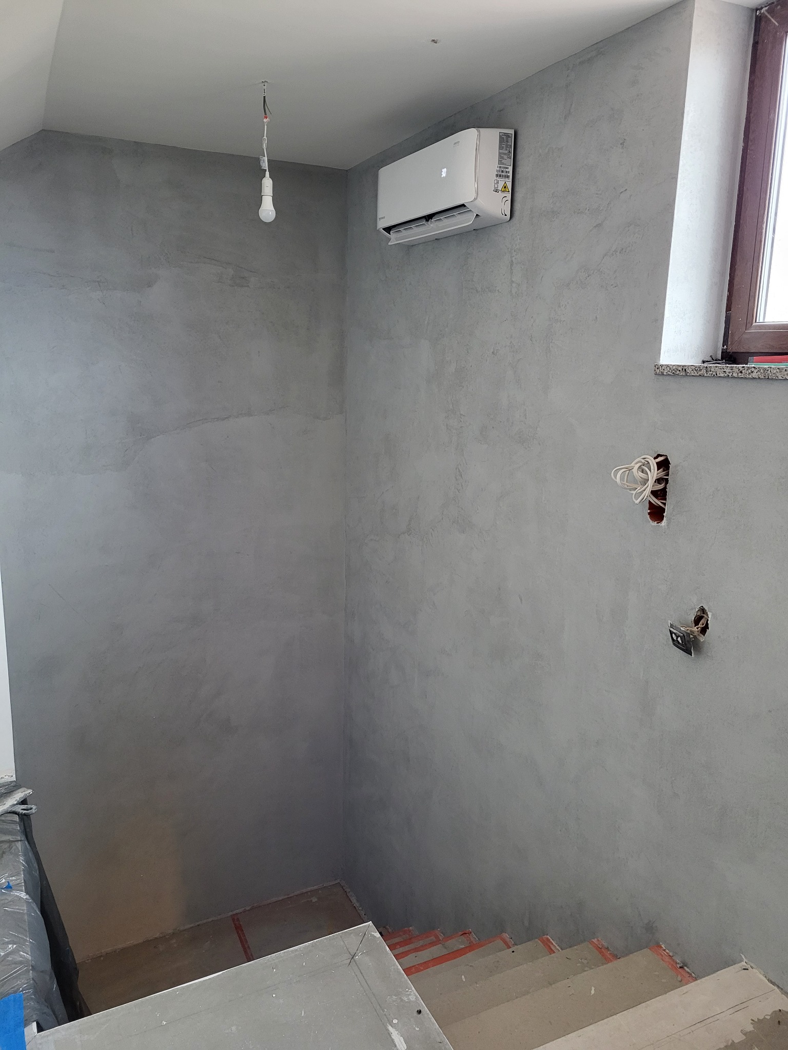 Wnętrze w trakcie remontu z zamontowaną klimatyzacją, widoczne niedokończone ściany z szarą fakturą, wystające kable elektryczne i schody w surowym stanie.