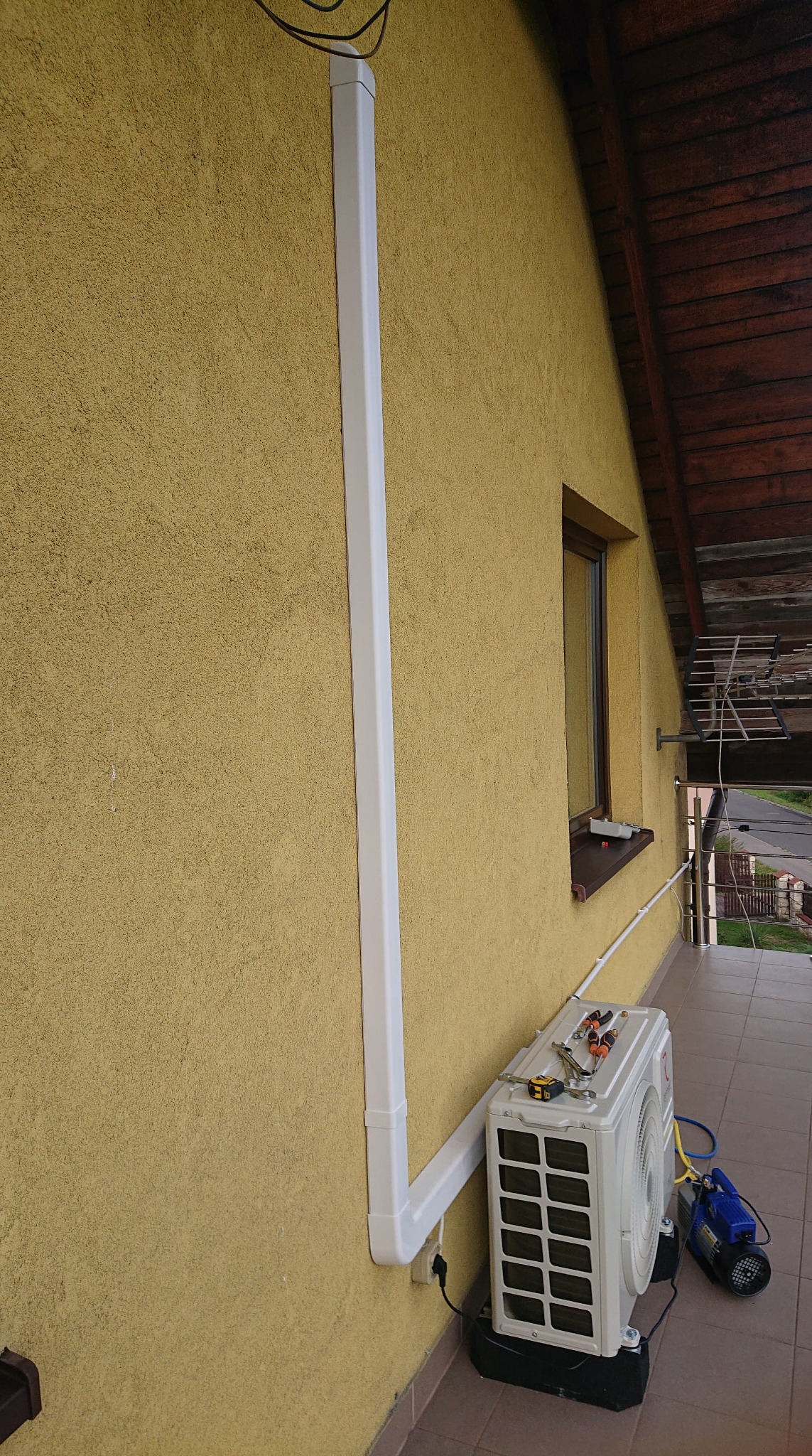 Zewnętrzna jednostka klimatyzacji zamontowana na elewacji domu, z widocznymi narzędziami na górze i prowadnicą kabli w kolorze białym.