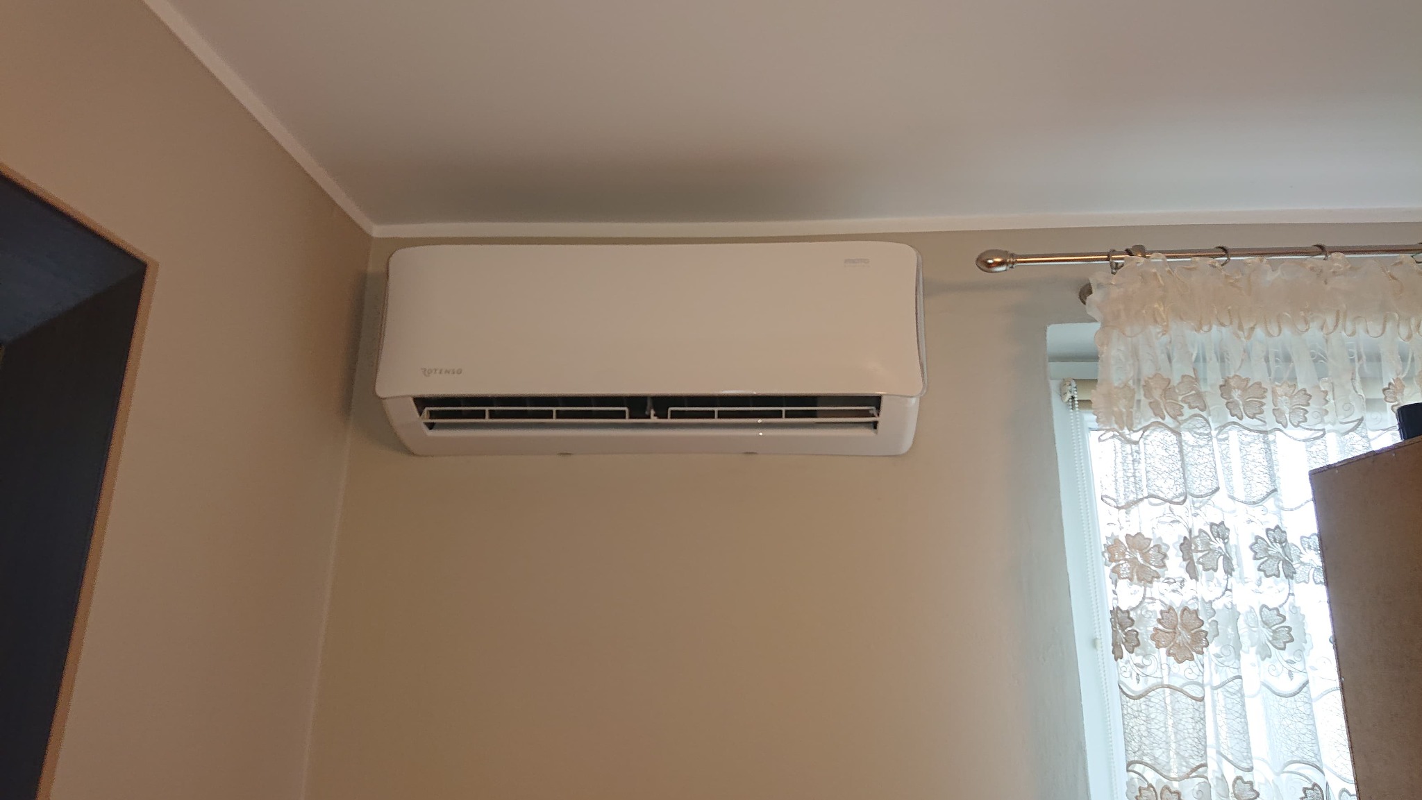 Biały klimatyzator marki Rotenso zamontowany na beżowej ścianie w pomieszczeniu, widoczne firanki w oknie.