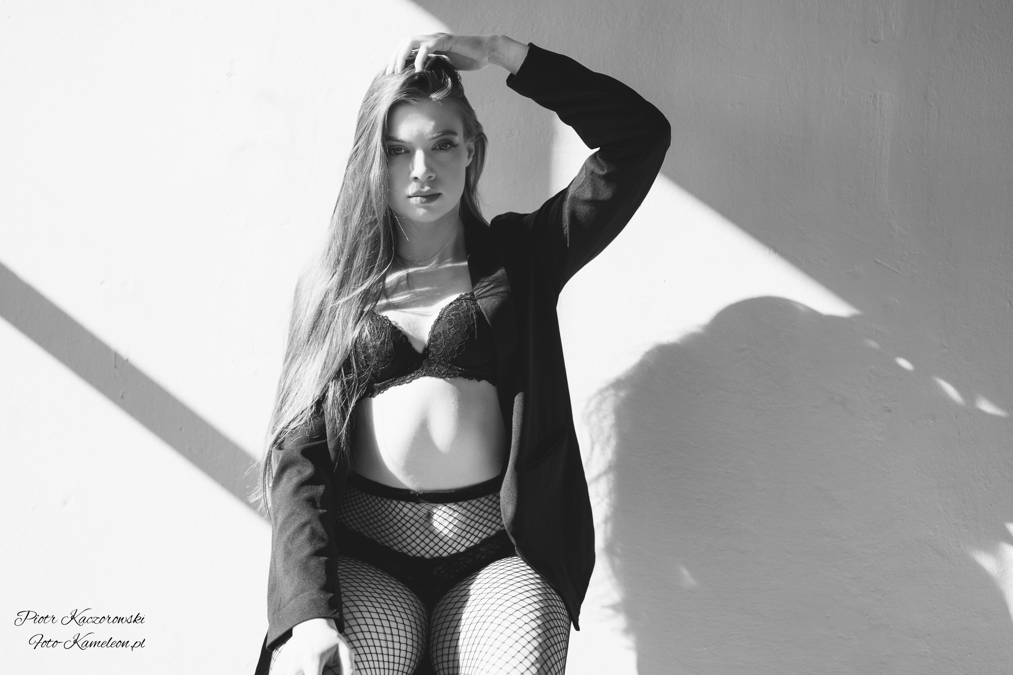 Portret kobiety w bieliźnie i marynarce, czarno-biała fotografia z mocnym światłem i cieniem na ścianie w tle.