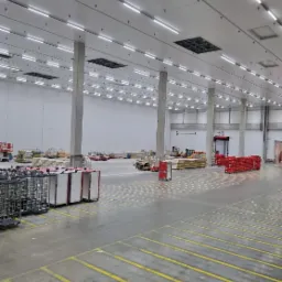 Wnętrze hali magazynowej z rzędami wózków, palet i materiałów budowlanych, oświetlone liniowymi lampami LED pod sufitem, widoczne betonowe słupy konstrukcyjne, żółte pasy na posadzce.