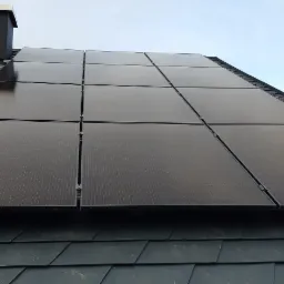Panele fotowoltaiczne zamontowane na ciemnym dachu z dachówki bitumicznej, widoczny komin w tle.