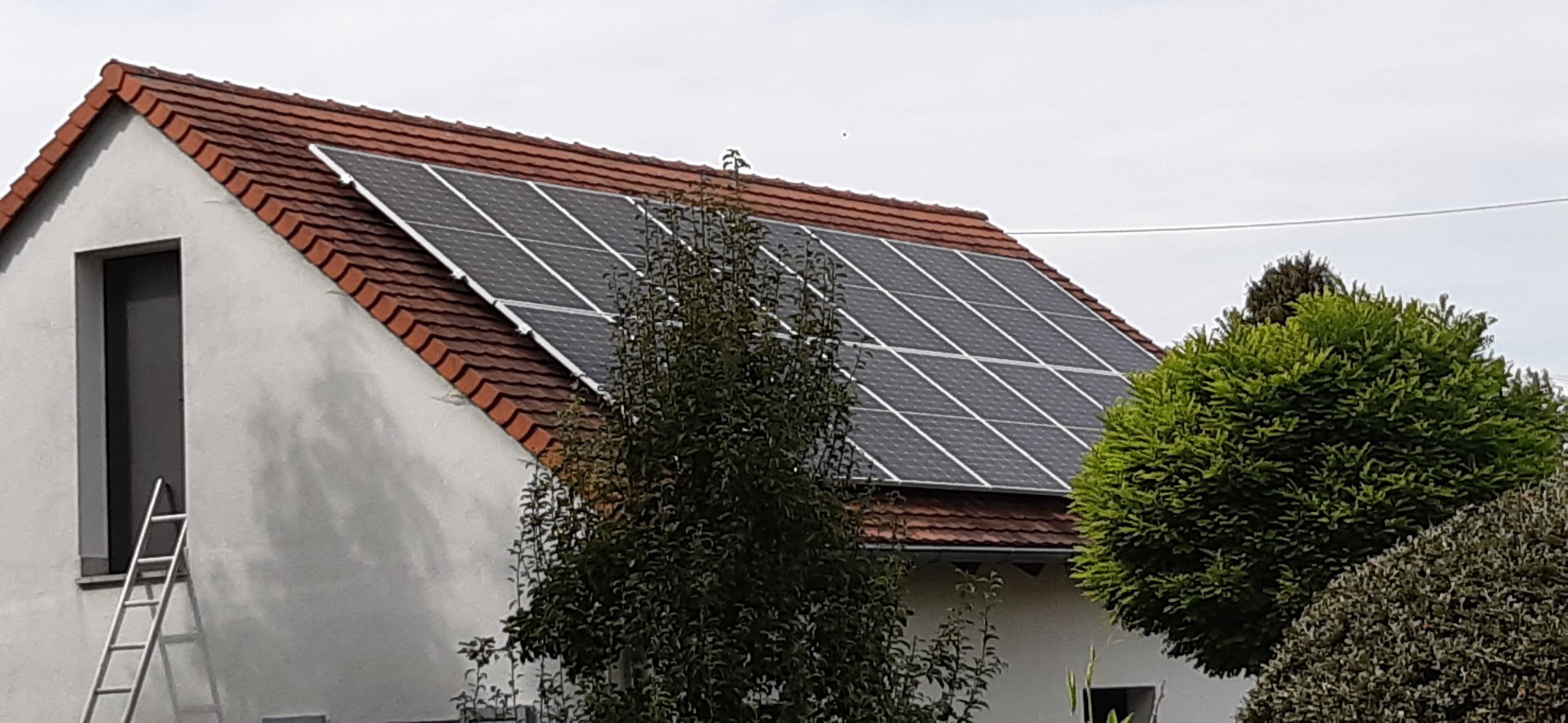 Panele słoneczne na dachu domu z czerwoną dachówką, widoczne z poziomu gruntu, z drabiną opartą o ścianę budynku.