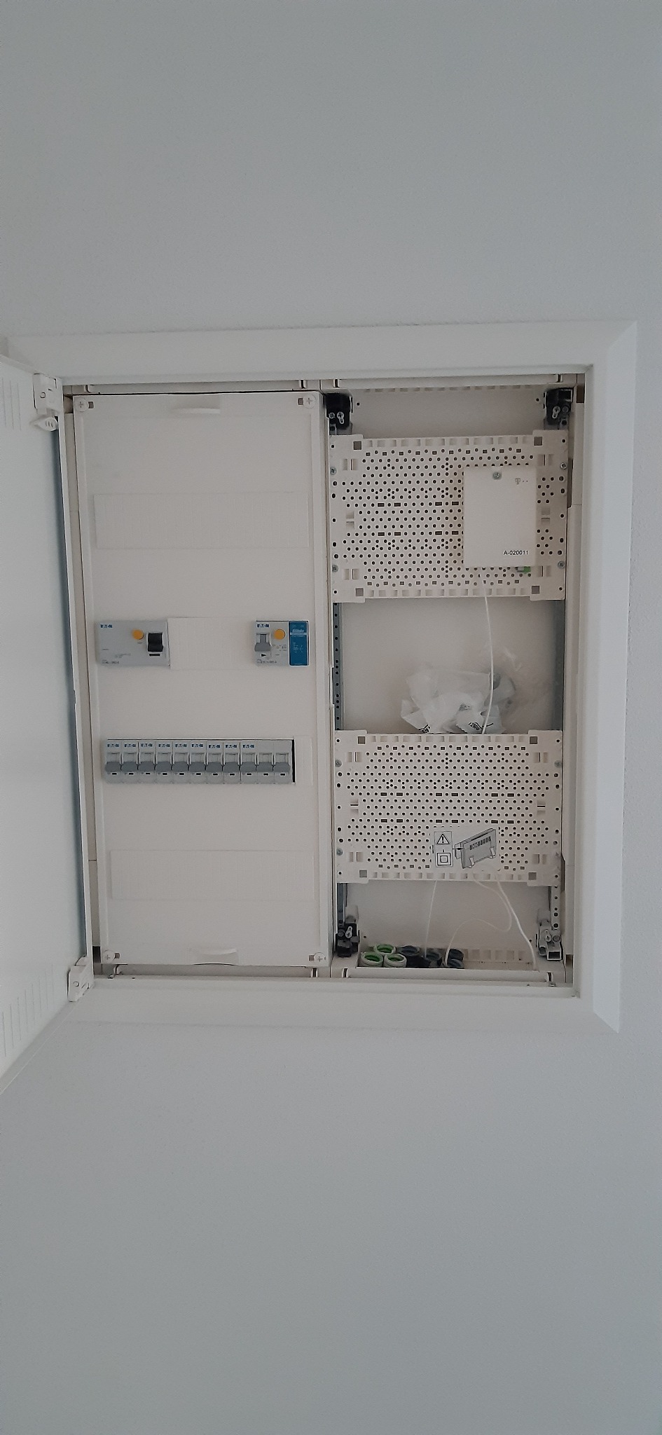 Otwarta skrzynka elektryczna w ścianie, widoczne bezpieczniki, wyłączniki i elementy montażowe, częściowo pusta przestrzeń wewnątrz.