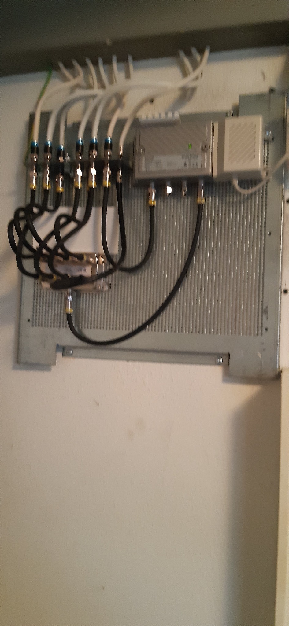 Szara metalowa płyta montażowa z podłączonymi kablami koncentrycznymi i rozdzielaczem sygnału, zamocowana na jasnej ścianie.