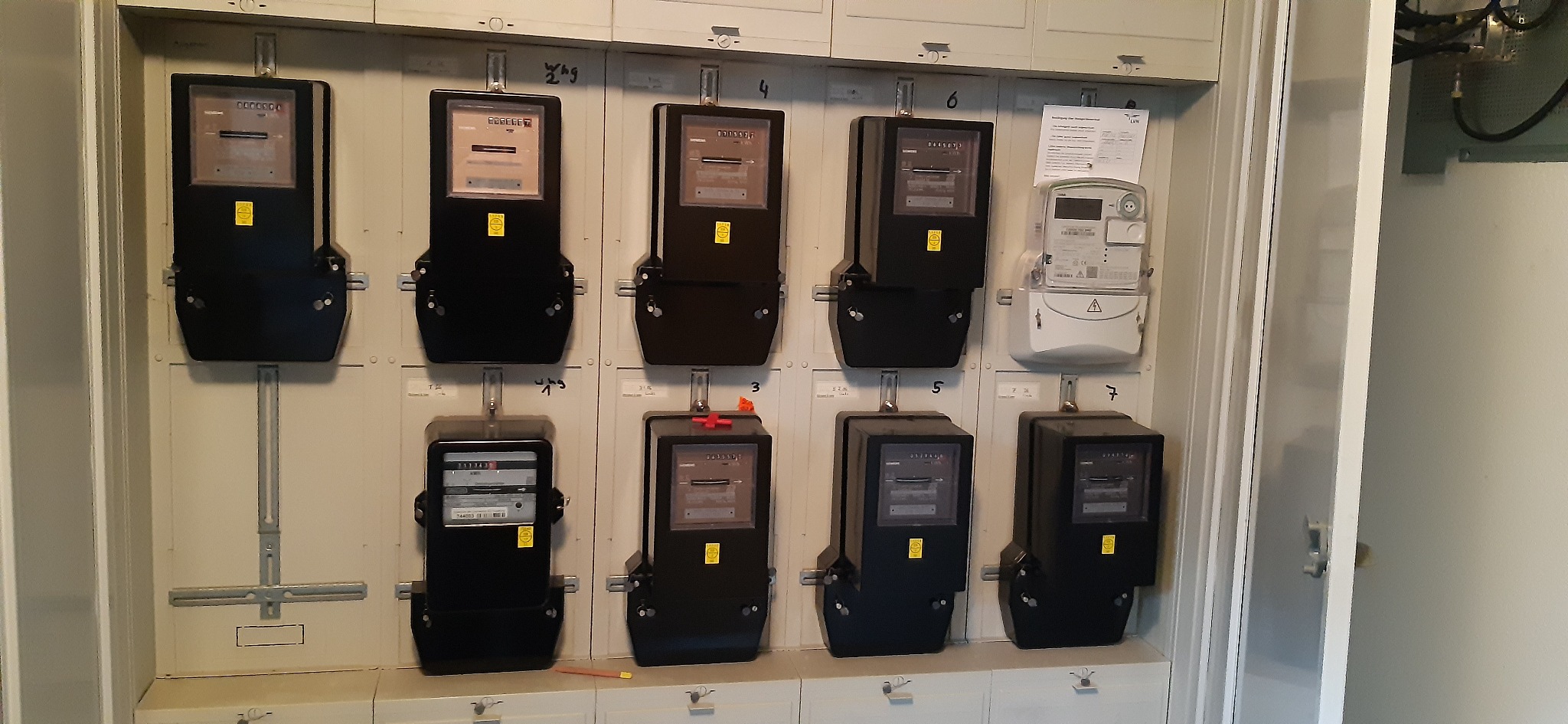 Rząd analogowych liczników energii elektrycznej w szafie rozdzielczej, z jednym nowszym, cyfrowym modelem po prawej stronie.