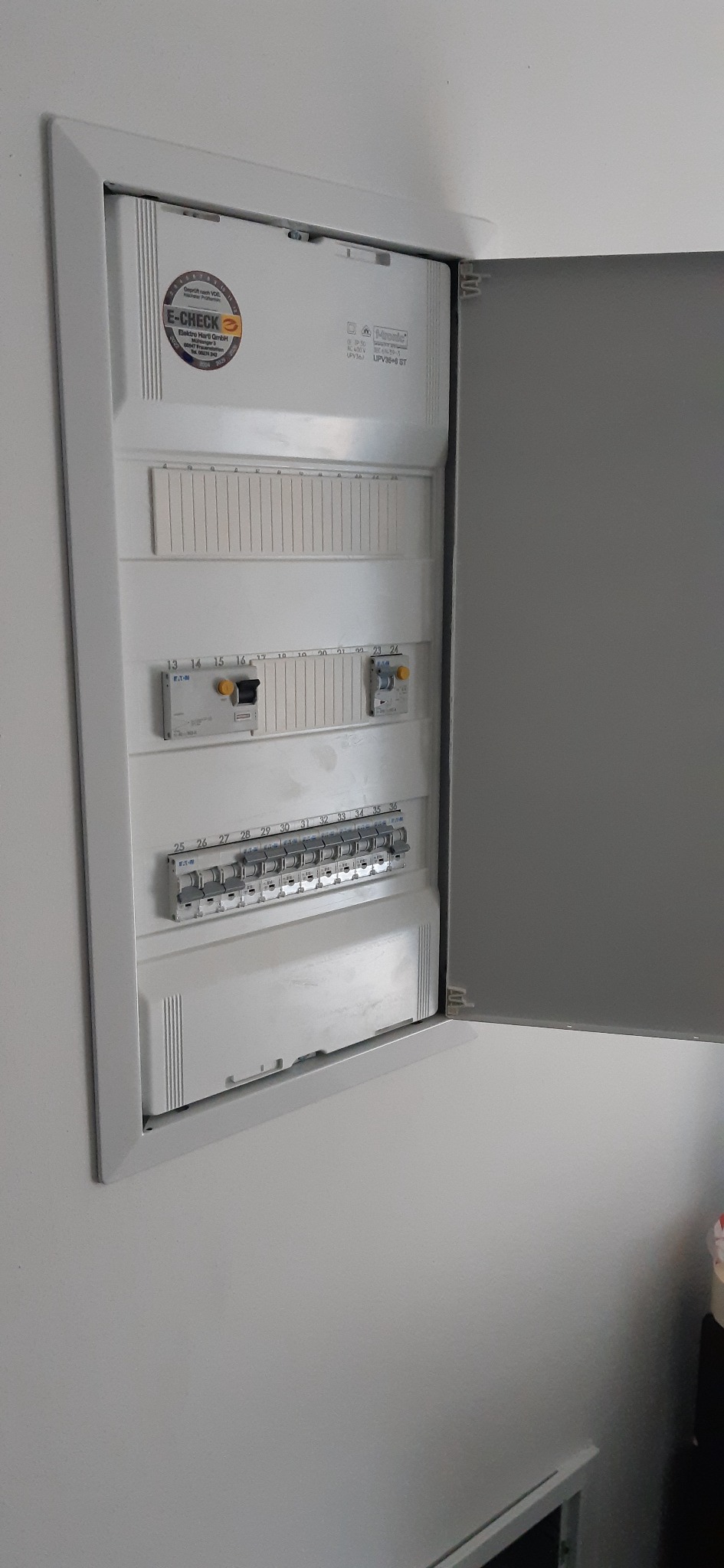 Otwarta skrzynka elektryczna z bezpiecznikami i wyłącznikami, widoczna naklejka certyfikacyjna E-CHECK na białej obudowie, szara ściana w tle.