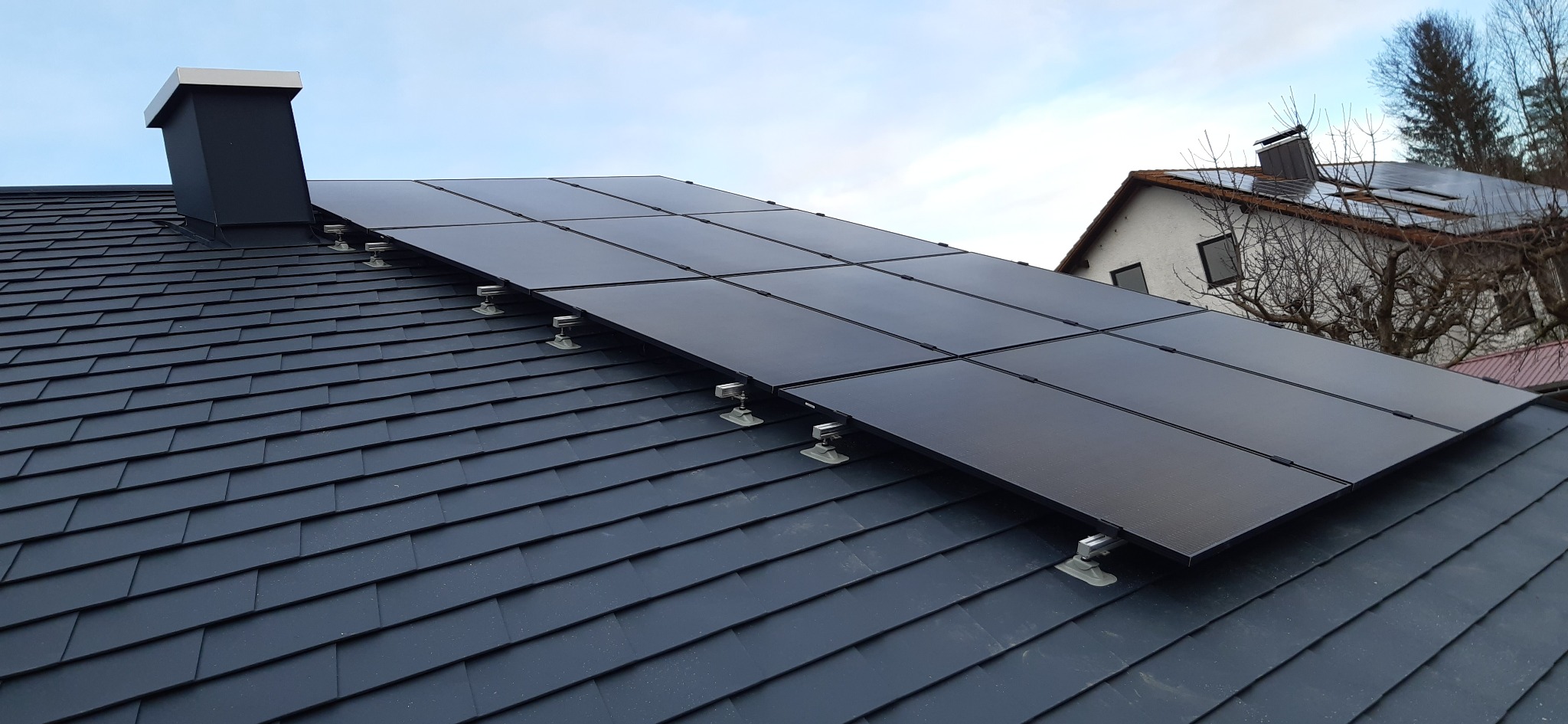 Czarne panele fotowoltaiczne zamontowane na ciemnoszarym dachu z dachówki ceramicznej, widoczny komin i fragment budynku w tle.
