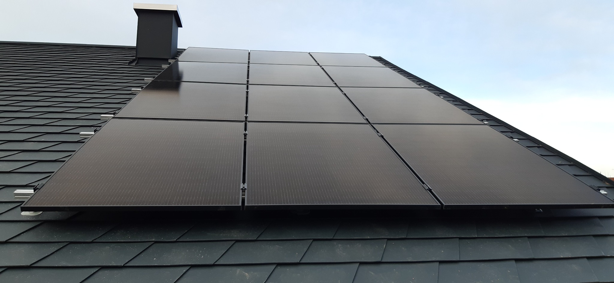 Panele fotowoltaiczne zamontowane na ciemnym dachu z dachówki bitumicznej, widoczny komin w tle.