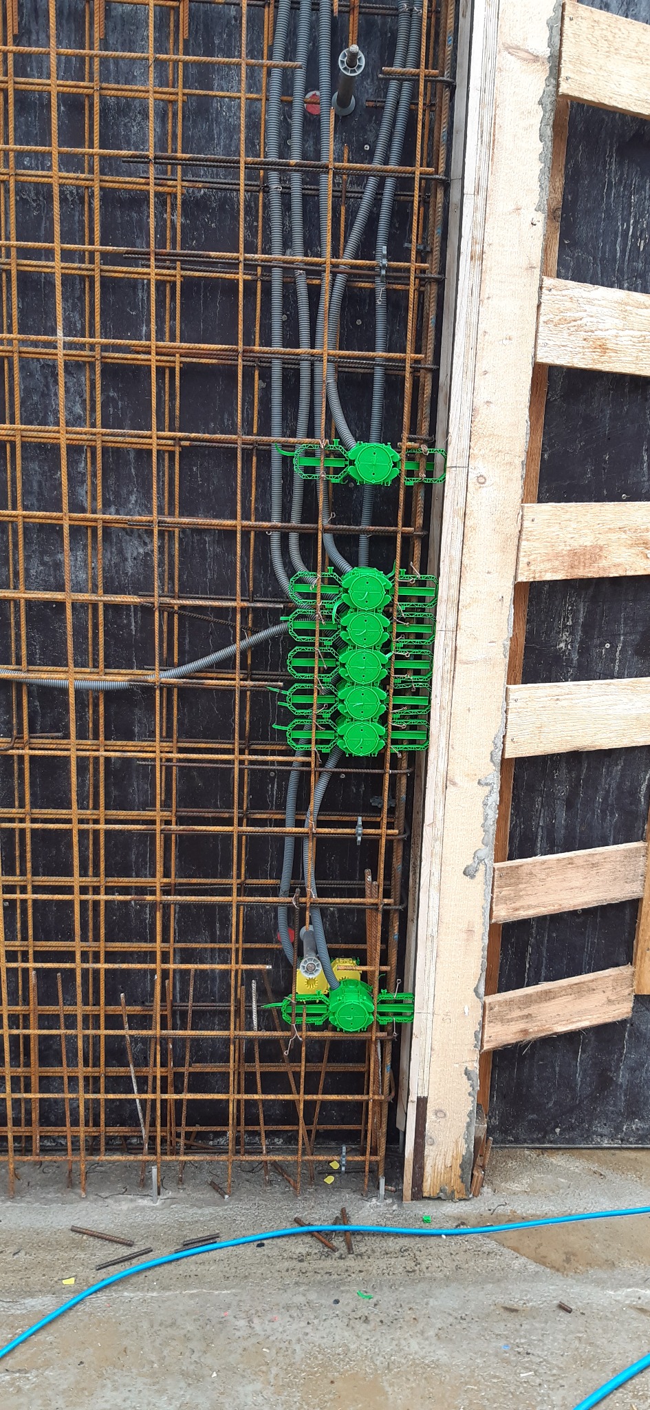 Zbrojenie ściany z zamontowanymi, zielonymi puszkami elektrycznymi i szarymi rurami instalacyjnymi, widoczne drewniane szalunki.