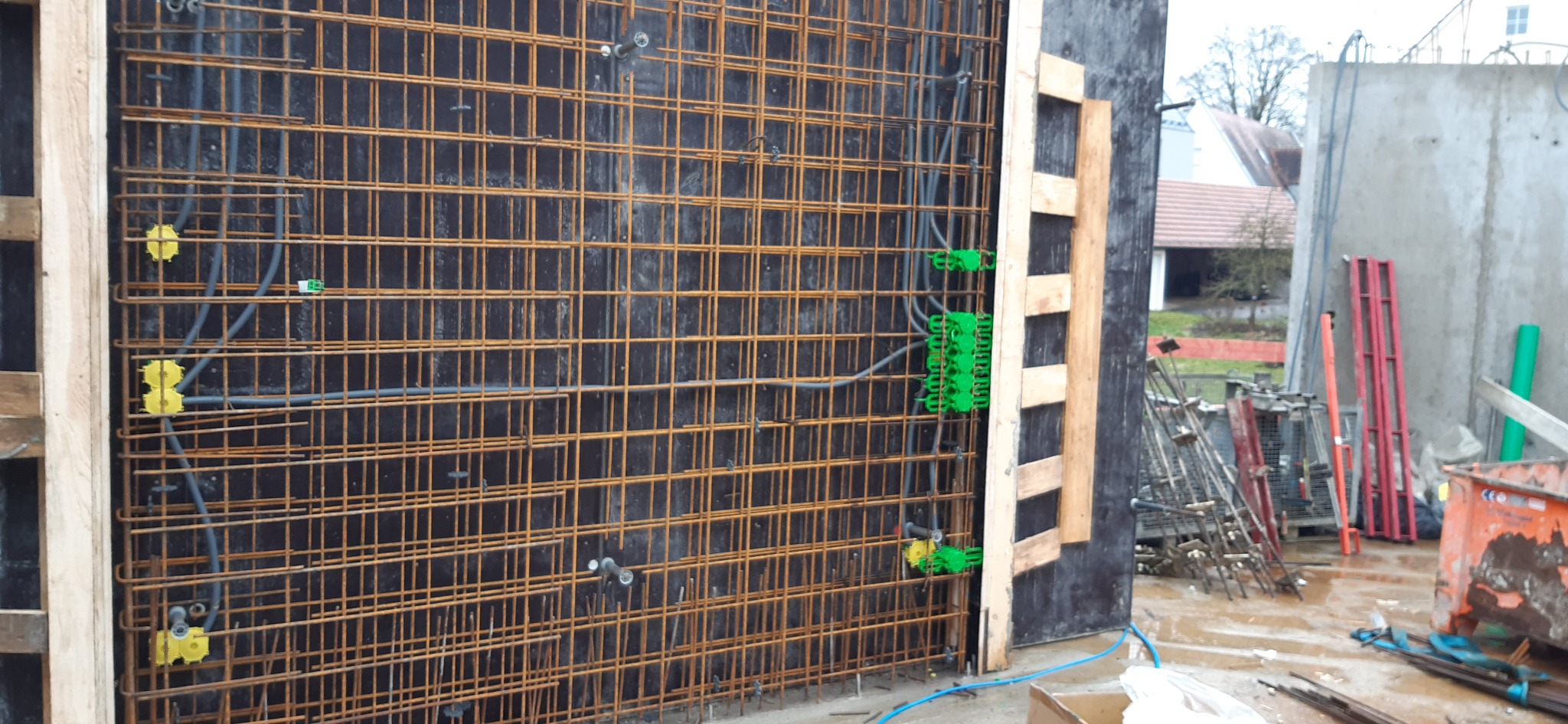 Zbrojenie ściany z widocznymi przewodami elektrycznymi zabezpieczonymi żółtymi i zielonymi puszkami, przygotowane do zalania betonem, na budowie w pochmurny dzień.