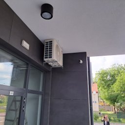 ICEKLIMA - Zewnętrzna jednostka klimatyzatora zamontowana na ścianie budynku komercyjnego, widoczna z dołu, obok okna z numerem U4A, z osobą rozmawiającą przez telefon w tle.