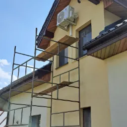 Montaż klimatyzacji na elewacji domu jednorodzinnego, widoczne rusztowanie i jednostka zewnętrzna klimatyzatora marki Gree na ścianie budynku z brązową stolarką okienną.